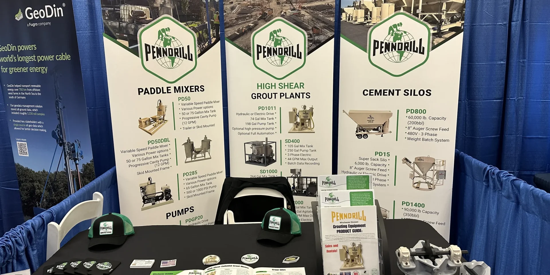 An exhibit booth with 3 white rollup banners and lots of stickers, brochures, and hats as giveaway items on the table.